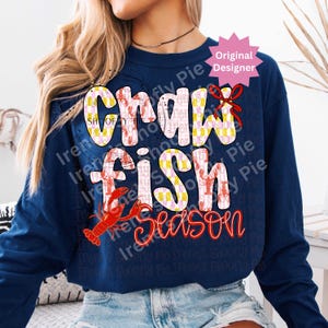 May include: Navy blue long-sleeve shirt with the text "Crawfish Season" in a playful font. The word "Crawfish" is in a checkered pattern with red crawfish illustrations. A red crawfish graphic is also featured.