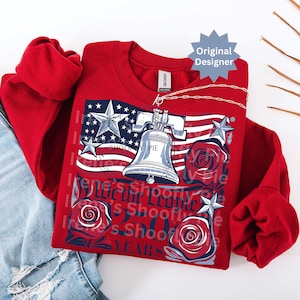 K&ouml;nnte beinhalten: Ein rotes Sweatshirt mit einem Design, das die amerikanische Flagge, eine Freiheitsglocke und Rosen zeigt. Der Text "Irene's Shoofly Pie" ist auf der Vorderseite aufgedruckt. Eine silberne Kette ist &uuml;ber dem Sweatshirt drapiert.