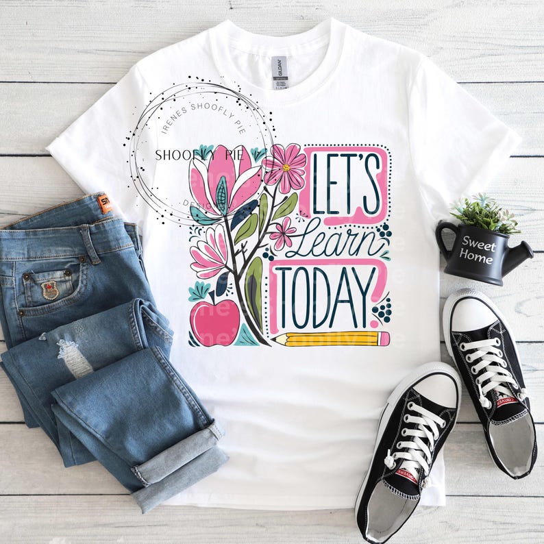 May include: White t-shirt with a colourful graphic design featuring flowers, an apple, a pencil, and the text "Let's Learn Today!"