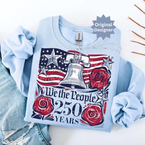 K&ouml;nnte beinhalten: Hellblaues Sweatshirt mit patriotischem Design. Es zeigt die amerikanische Flagge, die Freiheitsglocke und rote Rosen. Der Text lautet "We the People 250 Years". Das Design beinhaltet Sterne und Streifen.