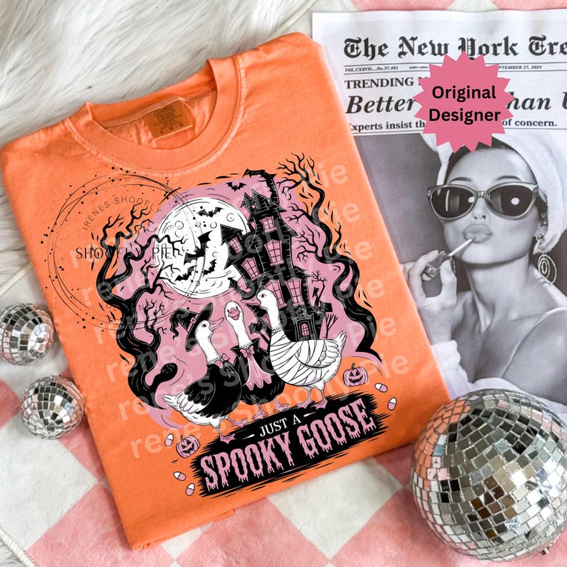 May include: Orange t-shirt with a Halloween-themed graphic. The design features a haunted house, bats, and three geese wearing witch hats, with the text "JUST A SPOOKY GOOSE". The shirt is displayed with disco balls and a newspaper.