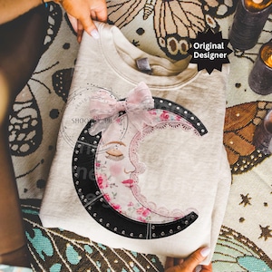 Puede incluir: Sudadera color crema con un diseño de media luna con una cara durmiente, estampado floral y ribete de encaje. La luna está delineada en negro con tachuelas plateadas. Se adjunta un lazo rosa en la parte superior. El texto "Original Designer" es visible.