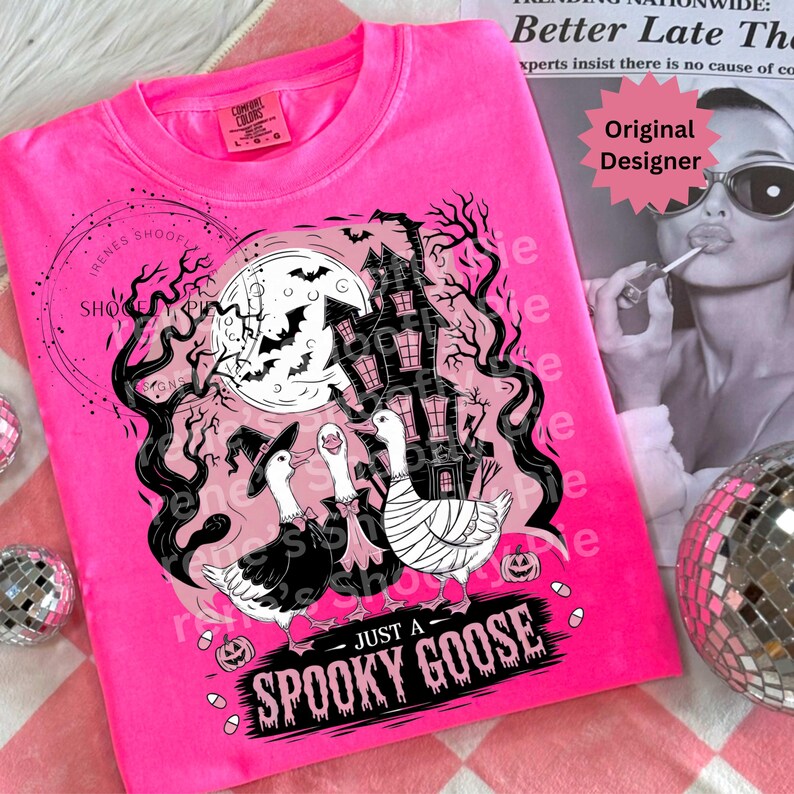 May include: A bright pink t-shirt featuring a Halloween-themed graphic. The design includes three geese in costumes, a haunted house, bats, and the text "JUST A SPOOKY GOOSE". The shirt has a crew neck and short sleeves.