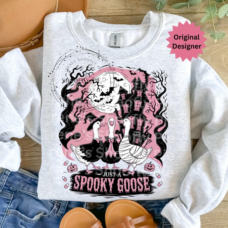 May include: A light gray sweatshirt featuring a Halloween-themed graphic. The design includes three geese dressed in costumes, a haunted house, a full moon, and the text "JUST A SPOOKY GOOSE".
