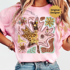 May include: Pink t-shirt featuring a large leaf design with floral and leopard print accents. The leaf is filled with various patterns and colors, including brown, yellow, and pink. The text "SHOOFY PIE" is visible on the shirt. The shirt has short sleeves.