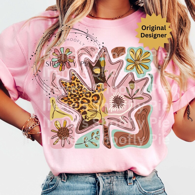 May include: Pink t-shirt featuring a large leaf design with floral and leopard print accents. The leaf is filled with various patterns and colors, including brown, yellow, and pink. The text "SHOOFY PIE" is visible on the shirt. The shirt has short sleeves.
