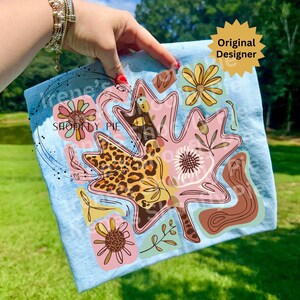 May include: A light blue decorative towel featuring a large pink leaf design with floral and leopard print accents. The towel has the text "SHOODY PIE" and "Original Designer" on it. The design includes various flowers and botanical elements.
