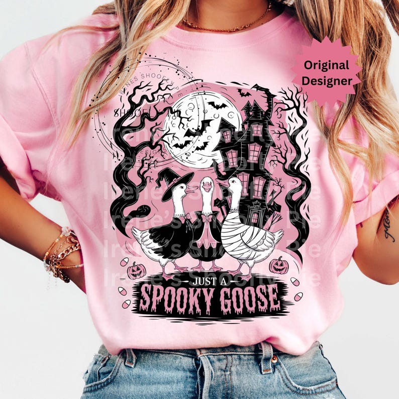 May include: Pink t-shirt featuring a Halloween-themed graphic. The design includes three geese dressed in costumes, a haunted house, bats, and the text "JUST A SPOOKY GOOSE". The design is in black and white.