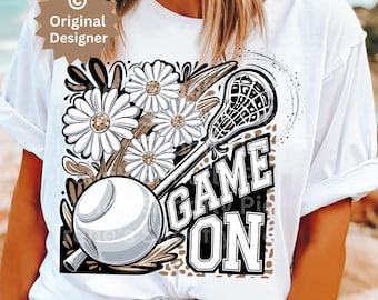 May include: White t-shirt with a graphic design featuring a lacrosse stick, flowers, and the text "GAME ON".