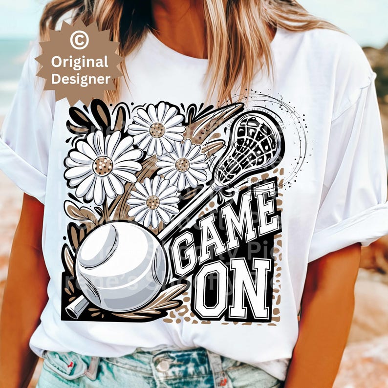 May include: White t-shirt with a graphic design featuring a lacrosse stick, flowers, and the text "GAME ON".