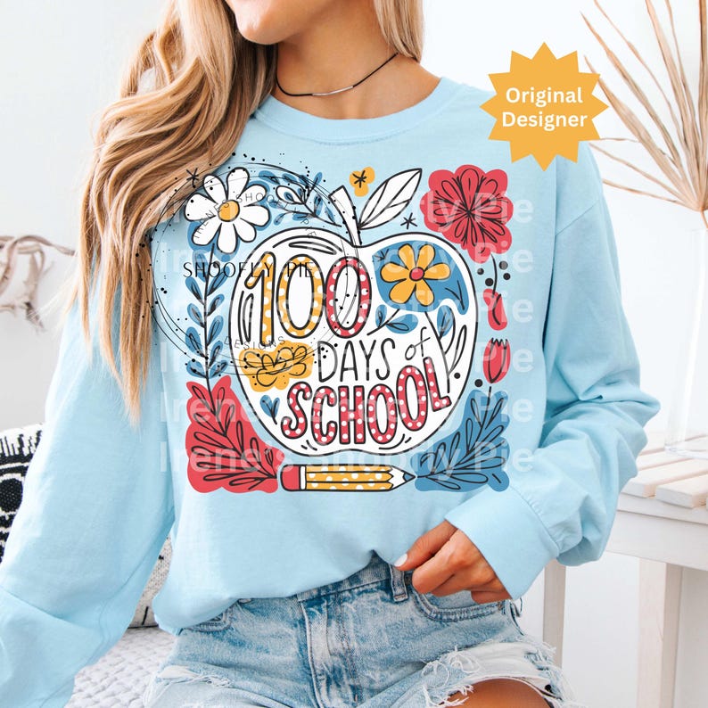 May include: Pale blue long-sleeved sweatshirt with a vibrant graphic. The design includes an apple shape with the text "100 Days of School", plus floral and pencil details. The graphic is in red, yellow, and white.