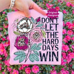 Op de afbeelding: Een lichtroze sweatshirt met een bloemenontwerp met luipaardprint. Het ontwerp bevat drie bloemen, bladeren en de tekst "DON'T LET THE HARD DAYS WIN" in het zwart. Het sweatshirt wordt door een hand omhoog gehouden.