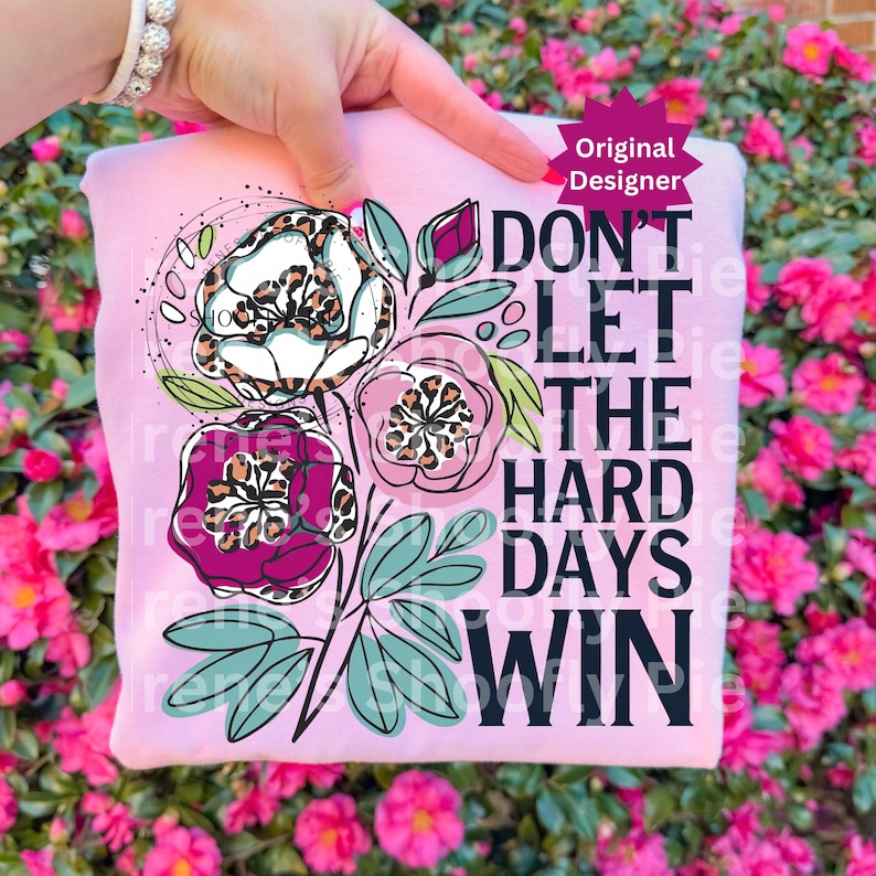 Op de afbeelding: Een lichtroze sweatshirt met een bloemenontwerp met luipaardprint. Het ontwerp bevat drie bloemen, bladeren en de tekst "DON'T LET THE HARD DAYS WIN" in het zwart. Het sweatshirt wordt door een hand omhoog gehouden.