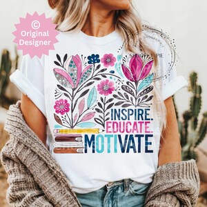 Op de afbeelding: Wit t-shirt met een bloemenontwerp met roze, blauwe en turquoise bloemen. Het shirt heeft de woorden "INSPIRE. EDUCATE. MOTIVATE." in een kleurrijk lettertype, samen met een potlood en boeken.