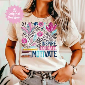 Op de afbeelding: Cr&egrave;mekleurig T-shirt met een bloemenontwerp met roze, blauwe en paarse bloemen. De woorden "INSPIRE. EDUCATE. MOTIVATE." staan onder een stapel boeken en een potlood. Het T-shirt heeft korte mouwen.