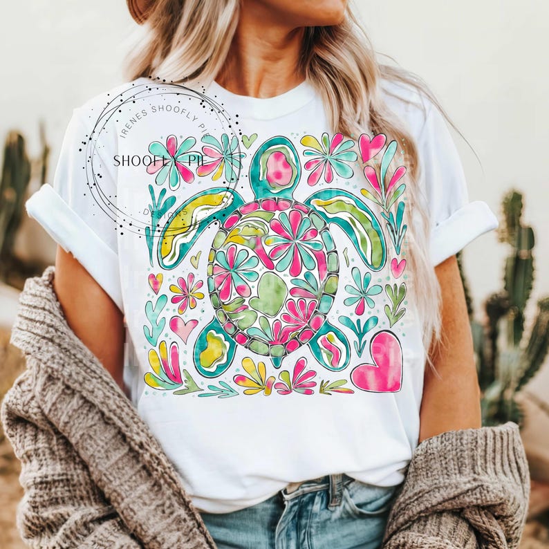 May include: A white t-shirt with a colorful floral design featuring a sea turtle. The turtle is surrounded by flowers, hearts, and swirls in shades of pink, blue, green, and yellow.