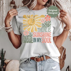May include: White t-shirt with a floral and sun design. The design includes the text "THERE IS SUNSHINE IN MY SOUL" in various colors. The shirt has short sleeves and a crew neck.