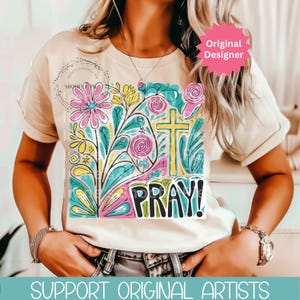 May include: Beige t-shirt featuring a colorful floral design with a cross and the word "PRAY!" in bold letters. The design includes pink, yellow, blue, and green elements. The shirt has short sleeves and a round neckline.