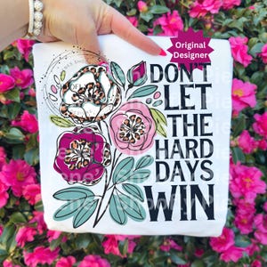 May include: White t-shirt with a floral design and the text "DON'T LET THE HARD DAYS WIN." The flowers have leopard print centers and are surrounded by green leaves. The words "Original Designer" are also visible.