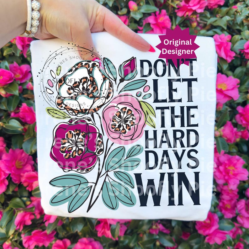 Op de afbeelding: Wit T-shirt met een bloemenontwerp en de tekst "DON'T LET THE HARD DAYS WIN." De bloemen hebben luipaardprintcentra en zijn omgeven door groene bladeren. De woorden "Original Designer" zijn ook zichtbaar.
