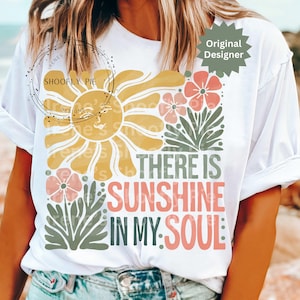 May include: White t-shirt with a colorful graphic design. The design features a yellow sun, pink flowers, and the text "THERE IS SUNSHINE IN MY SOUL" in coral and green. A casual, comfortable tee.