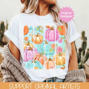 May include: White t-shirt featuring a colorful pumpkin design with floral accents. The pumpkins are in shades of pink, orange, and blue, set against a light blue background. The text "Original Designer" is visible. The bottom of the shirt reads "SUPPORT ORIGINAL ARTISTS."