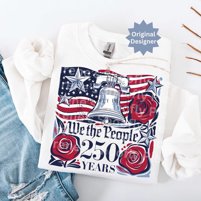 K&ouml;nnte beinhalten: Wei&szlig;es Sweatshirt mit einem patriotischen Design mit der amerikanischen Flagge, der Liberty Bell und Rosen. Der Text "We the People 250 Years" ist abgebildet. Eine silberne Halskette ist &uuml;ber dem Sweatshirt drapiert.