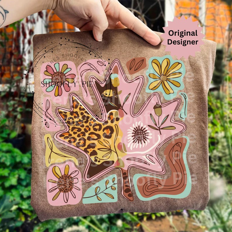 May include: A brown t-shirt featuring a large, stylized leaf design with a leopard print section, pink, brown, and yellow floral elements, and the text "SHOOFY PIE". The design includes an acorn and other botanical details.