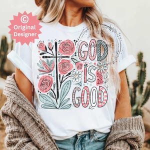 以下が含まれることがあります： 白いTシャツに花柄のデザインと「God is good」というピンクとブルーの文字が書かれています。