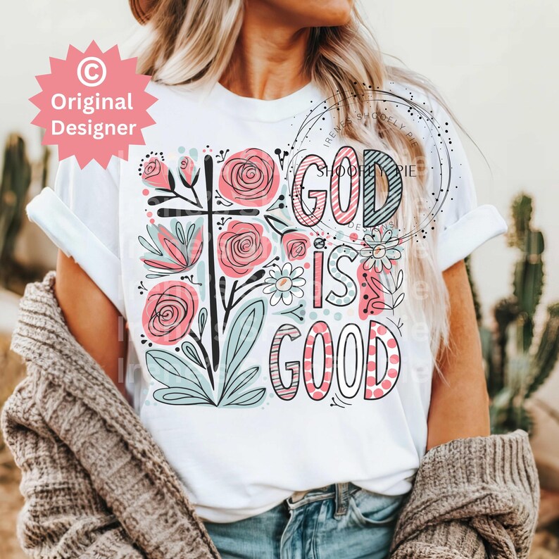 以下が含まれることがあります： 白いTシャツに花柄のデザインと「God is good」というピンクとブルーの文字が書かれています。