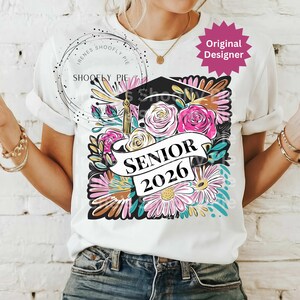 以下が含まれることがあります： 卒業キャップ、バラ、デイジーなどの花柄が特徴の白いTシャツ。「SENIOR 2026」の文字がバナーに表示されています。シャツは袖をまくり、丸いネックラインになっています。
