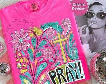 May include: A bright pink t-shirt with a colorful floral and cross design. The design includes the word "PRAY!" in bold letters. The shirt also features the text "Original Designer" and "IRENES SHOOFLY PIE".