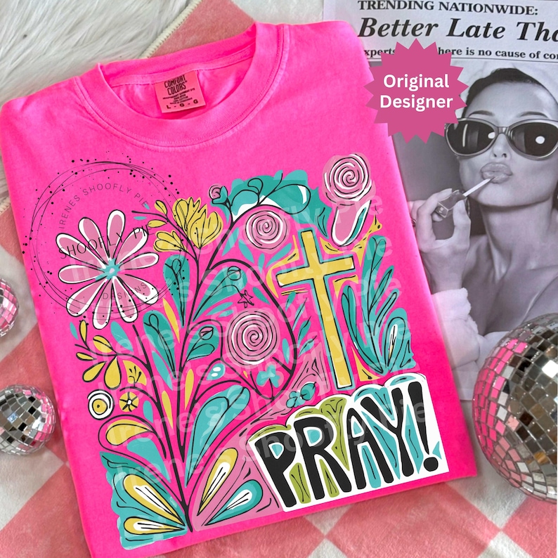 May include: A bright pink t-shirt with a colorful floral and cross design. The design includes the word "PRAY!" in bold letters. The shirt also features the text "Original Designer" and "IRENES SHOOFLY PIE".