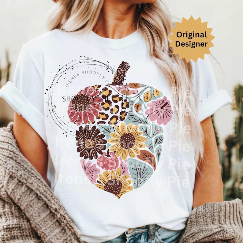 May include: White t-shirt featuring a floral pumpkin design. The pumpkin is composed of various flowers, leopard print, and leaves in shades of pink, yellow, and brown. The text "Irenes Shoofly Pie" is printed on the shirt.