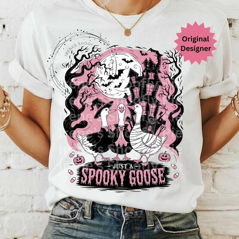 May include: White t-shirt featuring a Halloween-themed graphic. The design includes three geese dressed in costumes, a haunted house, bats, and the text "Just a Spooky Goose". The design is in black and pink.