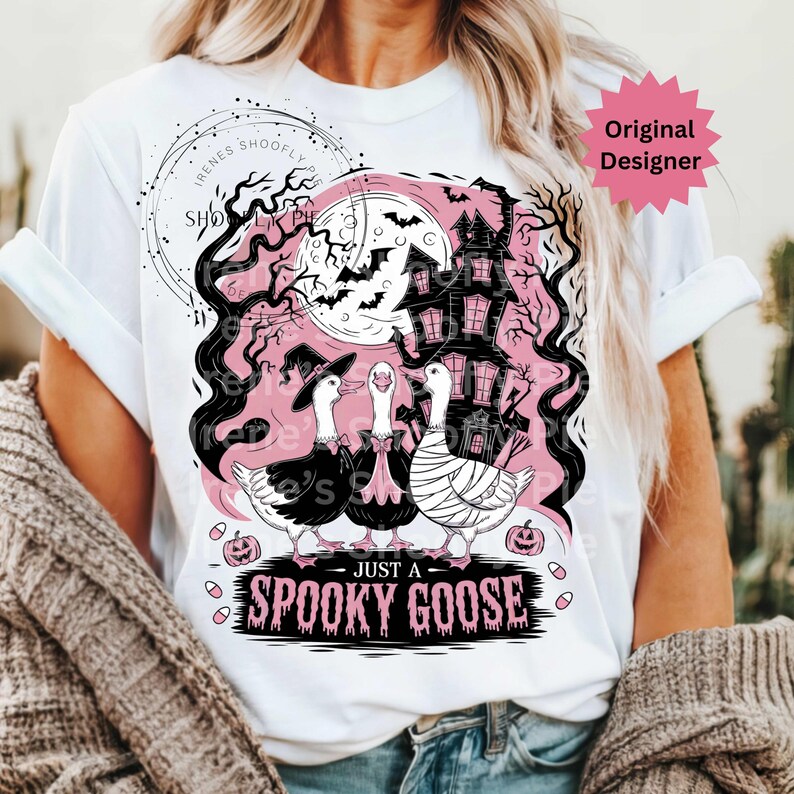 May include: White t-shirt featuring a Halloween-themed graphic. The design includes three geese dressed in costumes, a haunted house, bats, and the text "JUST A SPOOKY GOOSE". The design is in black and pink.