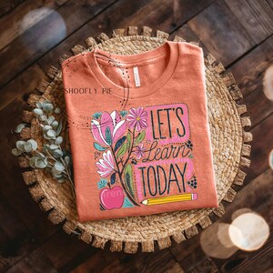 May include: A peach coloured t-shirt with a floral design and the text "Let's Learn Today" in a colourful font. A yellow pencil and a red apple are also included in the design.