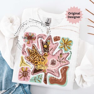 May include: White sweatshirt featuring a colorful leaf design with floral and leopard print accents. The design includes pink, yellow, brown, and teal elements. The text "Irenes Shoofly Pie" is printed on the sweatshirt.