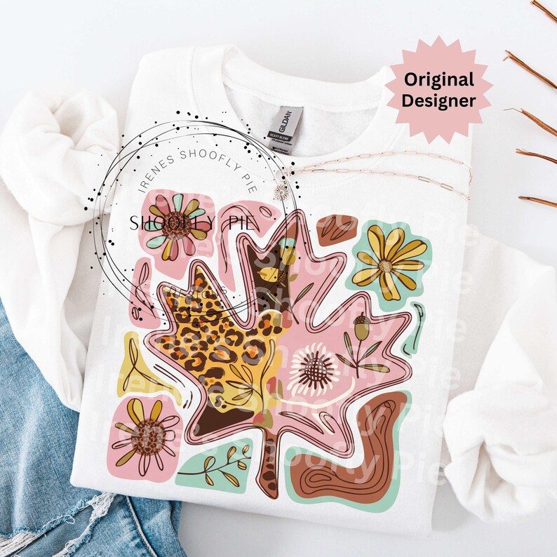 May include: White sweatshirt featuring a colorful leaf design with floral and leopard print accents. The design includes pink, yellow, brown, and teal elements. The text "Irenes Shoofly Pie" is printed on the sweatshirt.