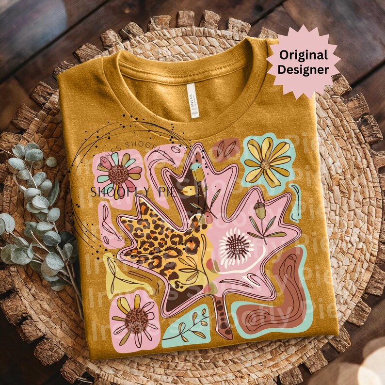 May include: Mustard yellow t-shirt featuring a large leaf design with floral and leopard print accents. The leaf is pink and brown, with yellow and teal floral elements. The shirt has the text "Original Designer" on a starburst.