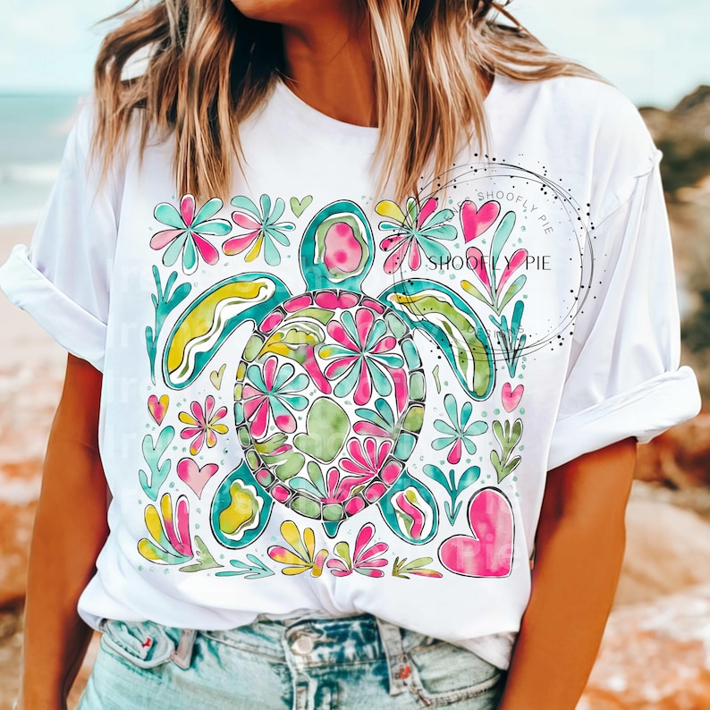 May include: White t-shirt with a colorful, abstract design of a sea turtle surrounded by flowers and hearts. The text "Shooly Pie" is printed on the shirt.