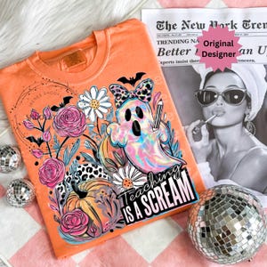 May include: Orange t-shirt with a colorful ghost graphic holding a pencil, surrounded by flowers, bats, and pumpkins. The text "Teaching is a Scream" is displayed on the front. A fun, Halloween-themed tee.