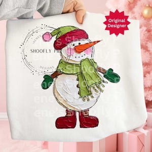 Op de afbeelding: Witte sweatshirt met een geschilderd sneeuwmanontwerp. De sneeuwman heeft een wortelneus, roze wangen en draagt een groene sjaal, hoed en wanten. Het ontwerp bevat de tekst "SHOOFY PIE DESIGNS" en een rode ster met "Original Designer".