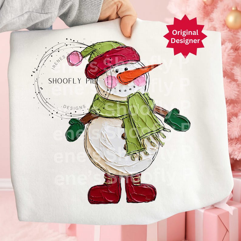 Op de afbeelding: Witte sweatshirt met een geschilderd sneeuwmanontwerp. De sneeuwman heeft een wortelneus, roze wangen en draagt een groene sjaal, hoed en wanten. Het ontwerp bevat de tekst "SHOOFY PIE DESIGNS" en een rode ster met "Original Designer".