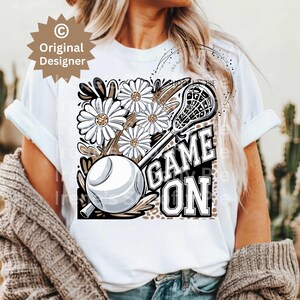 May include: White t-shirt with a brown and white lacrosse stick, ball, and flowers. The text "GAME ON" is printed in black letters.