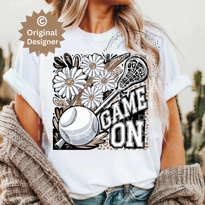 May include: White t-shirt with a brown and white lacrosse stick, ball, and flowers. The text "GAME ON" is printed in black letters.