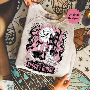 May include: A light beige sweatshirt featuring a Halloween-themed graphic. The design includes three geese in costumes, a haunted house, bats, and the text "Just a Spooky Goose". The design is in black and pink.