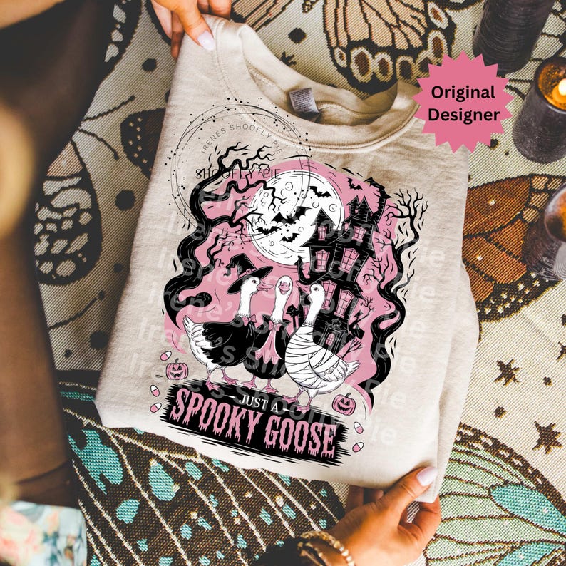 May include: A light beige sweatshirt featuring a Halloween-themed graphic. The design includes three geese in costumes, a haunted house, bats, and the text "Just a Spooky Goose". The design is in black and pink.