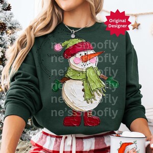 Op de afbeelding: Een donkergroene sweatshirt met een grillig sneeuwmanontwerp. De sneeuwman heeft een rode hoed, groene sjaal en wanten en rode laarzen. Het ontwerp staat tegen een donkergroene achtergrond. De woorden "Original Designer" staan in een rode ster.