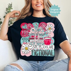 May include: Black t-shirt with a colorful graphic design featuring a stack of books tied with a pink bow, roses, daisies, and a coffee cup. The text "MY PLANS ARE FICTIONAL" is displayed.